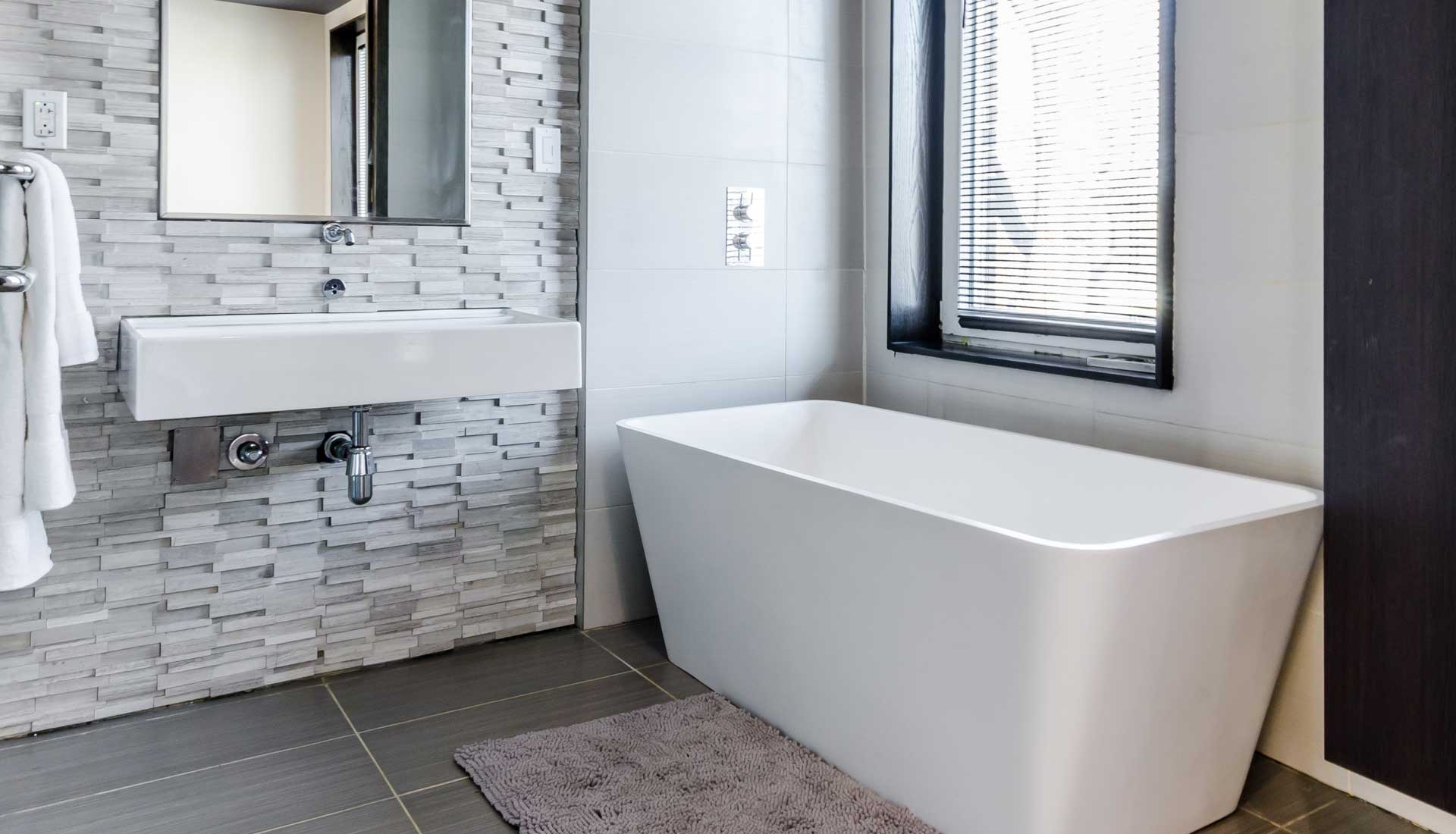 White bathub in bathroom under window with white blinds sits beside modern white sink with a grey tile backsplash